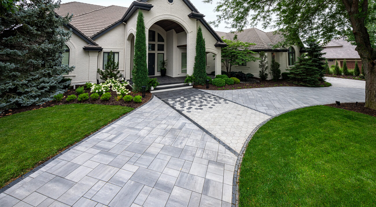 Modern paver walkway with geometric patterns leading to elegant home entrance, bordered by manicured landscaping and greenery.