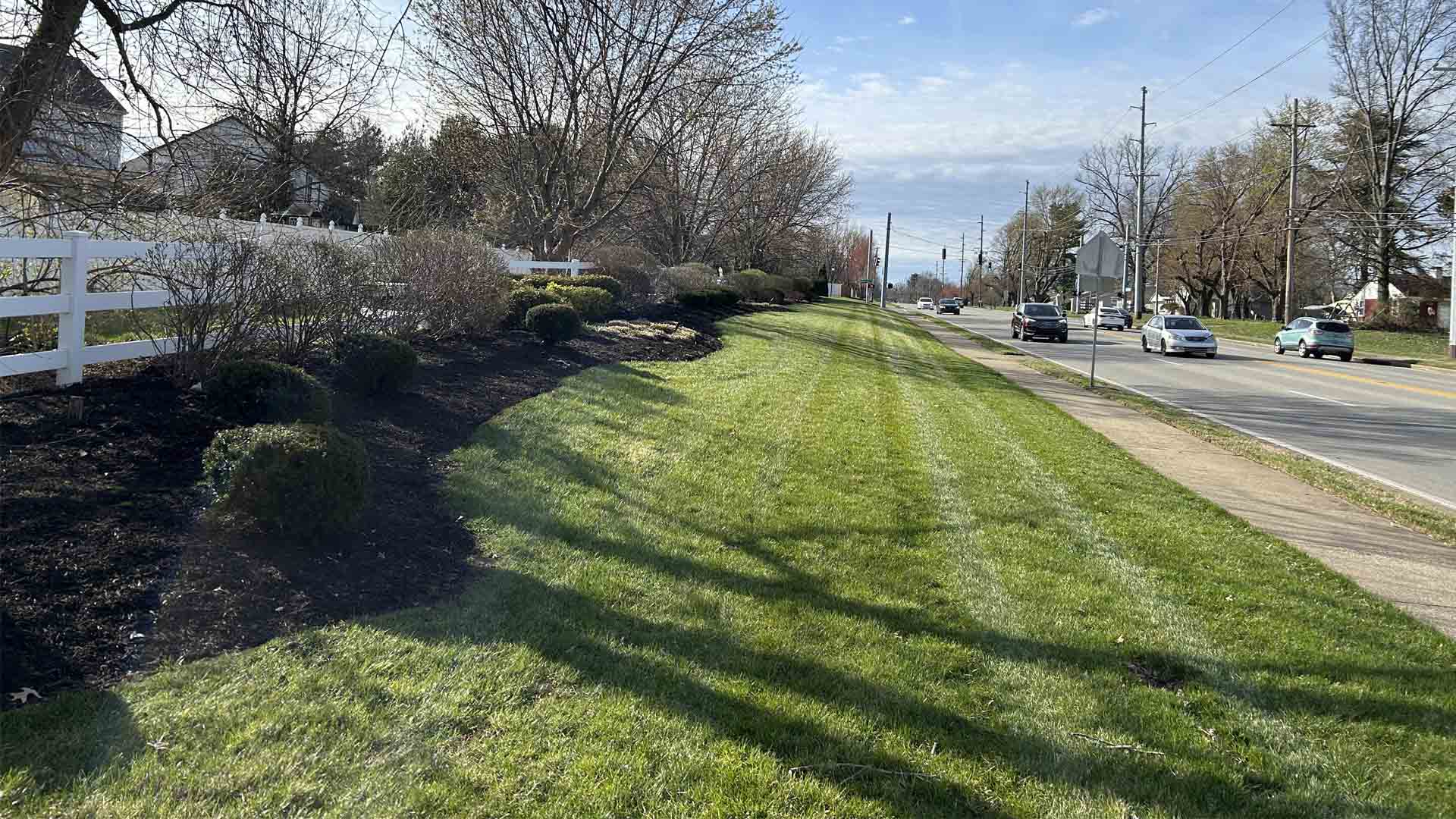 Beautifully manicured lawn beside roadway, featuring fresh mulch, manicured shrubs, and passing vehicles under daylight.