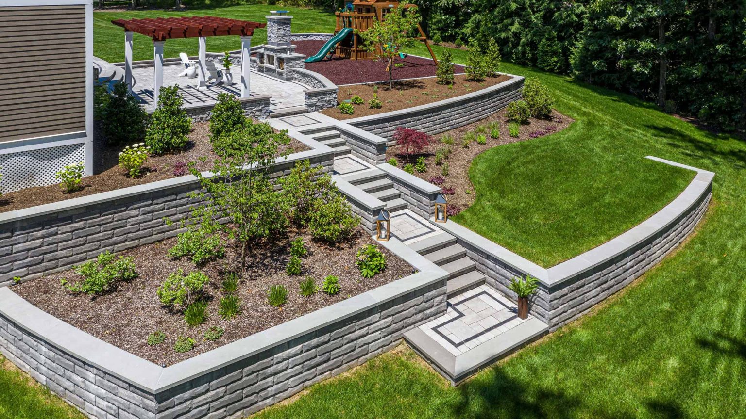 Landscape design with tiered retaining walls, stone walkways, patio with outdoor seating, and playground.