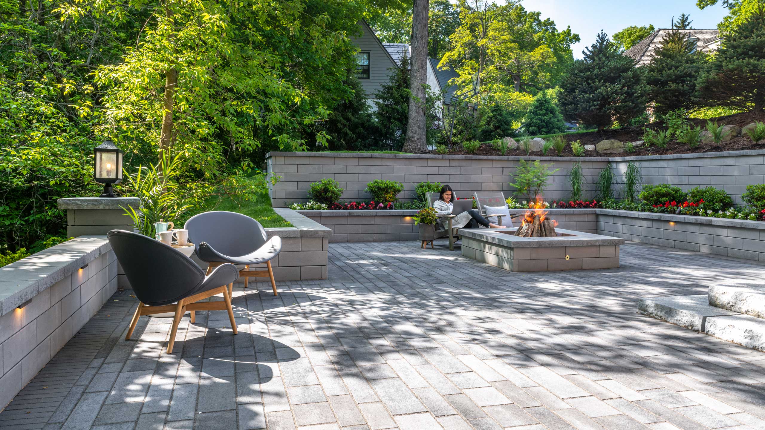 Outdoor living space with gray brick patio, fire pit, seating, and retaining wall with plants.